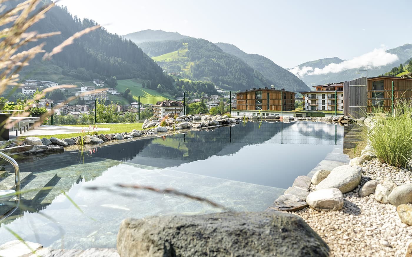 Ein Pool mit Steinen und Pflanzen vor einer Bergkulisse und modernen Geb&auml;uden