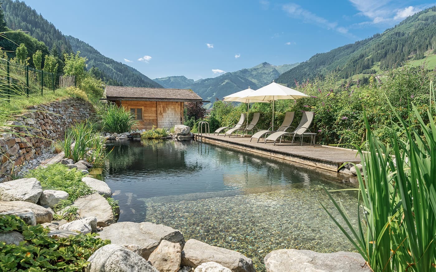 Naturteich mit Liegest&uuml;hlen und Sonnenschirmen, umgeben von Bergen und gr&uuml;ner Vegetation