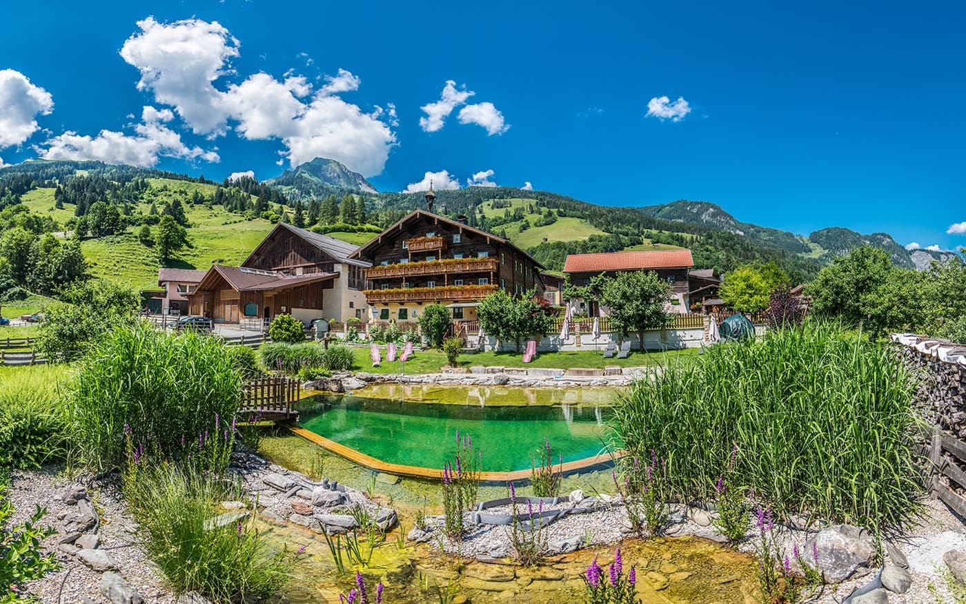 Ein malerisches Bauernhaus vor gr&uuml;nen Bergen und blauem Himmel mit einem kleinen Teich im Vordergrund