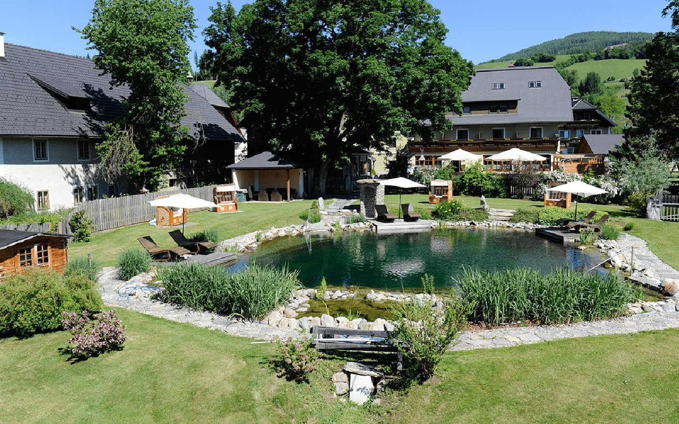Garten mit Teich, Sonnenliegen und Sonnenschirmen vor einem Geb&auml;ude in gr&uuml;ner Landschaft