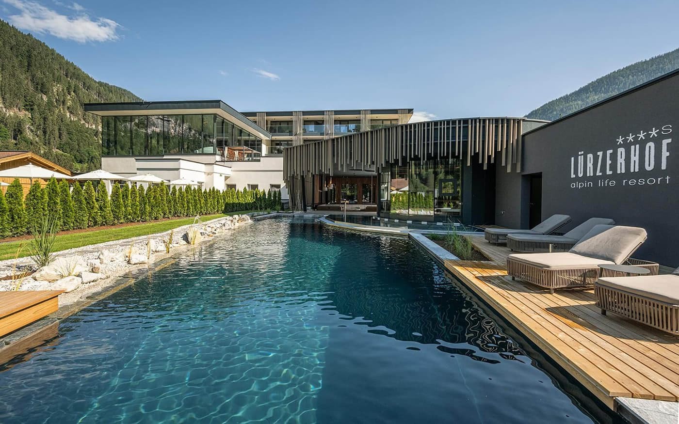 Luxuri&ouml;ser Outdoor-Pool vor einem modernen Alpenresort mit Sonnenliegen und Bergkulisse
