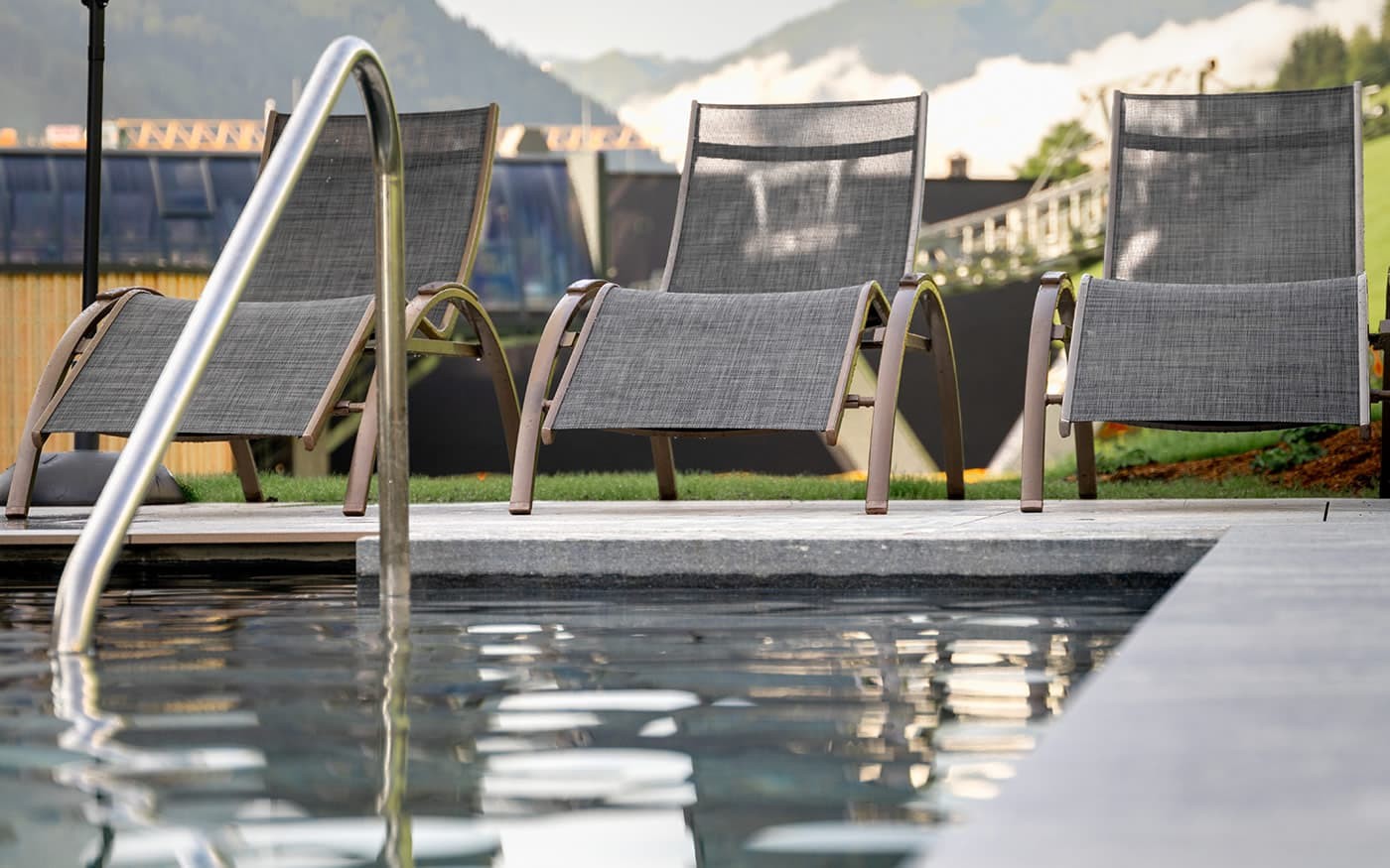 Drei Liegest&uuml;hle stehen neben einem Pool mit Bergkulisse im Hintergrund
